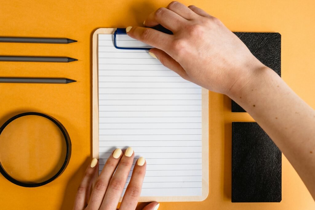 pexels-photo-7718883-7718883 Top view of hands on a notepad with various stationery items on a yellow desk background.